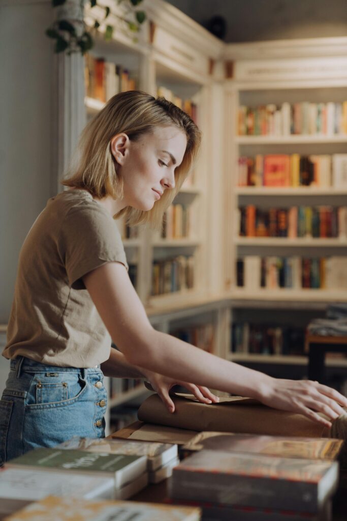 Une libraire est en train d'emballer un livre pour faire un joli papier cadeau. En arrière-plan, on peut voir des rangés de livres alignés dans des grandes bibliothèques