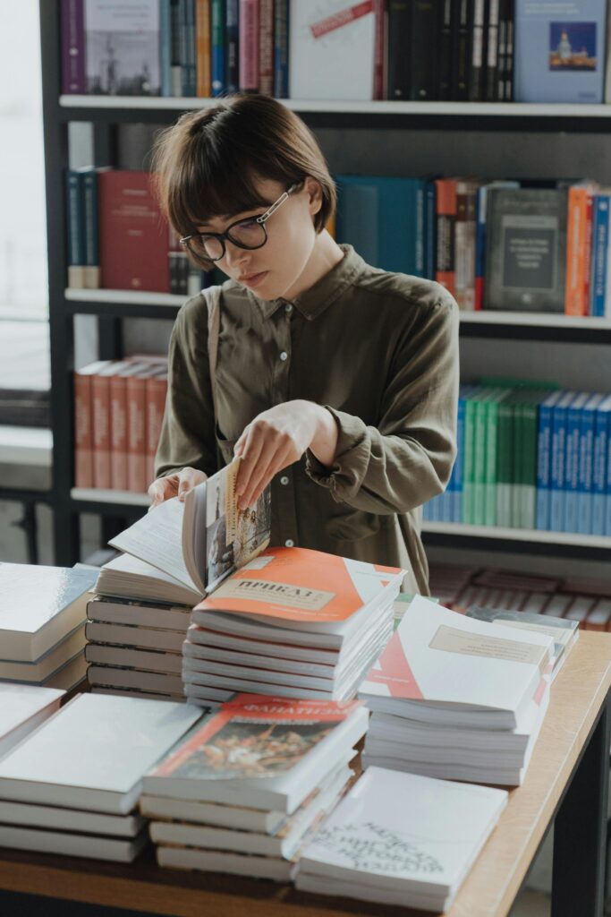 Une jeune femme feuillette un livre qui se trouve sur table en librairie. Derrière elle, on peut voir une grande bibliothèque avec des livres bien alignés
