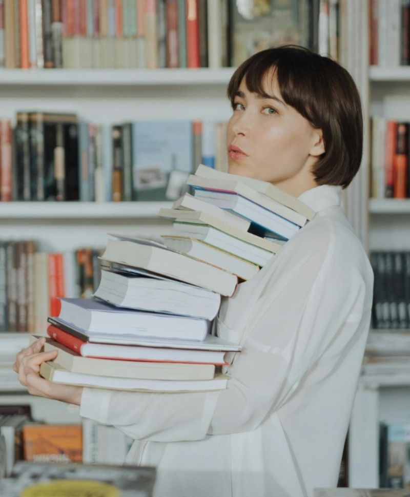 Une jeune femme tenant de nombreux livres dans les mains, qui lui montent jusqu'au menton. Elle se trouve dans une librairie.